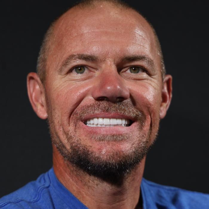 A man with a shaved head and a neatly trimmed beard smiles at the camera, wearing a blue shirt against a dark background.