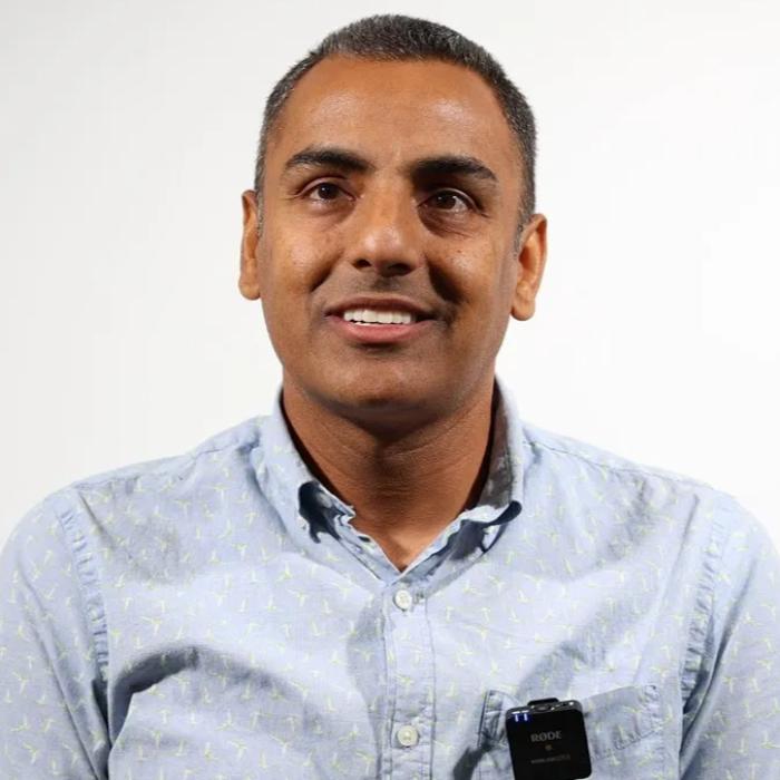 A man wearing a light blue button-up shirt with a Rode microphone attached to it, smiling against a plain white background.