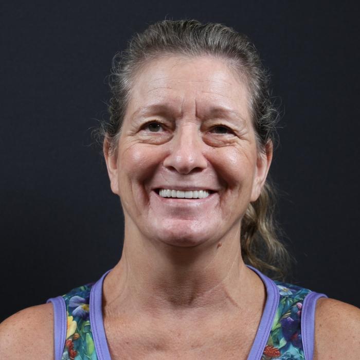 A middle-aged woman with a ponytail smiles at the camera. She wears a floral-patterned sleeveless top and is posed against a dark background.