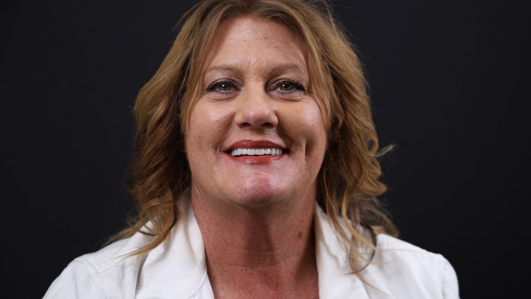 A woman with shoulder-length blond hair smiles at the camera, wearing a white collared shirt against a black background.