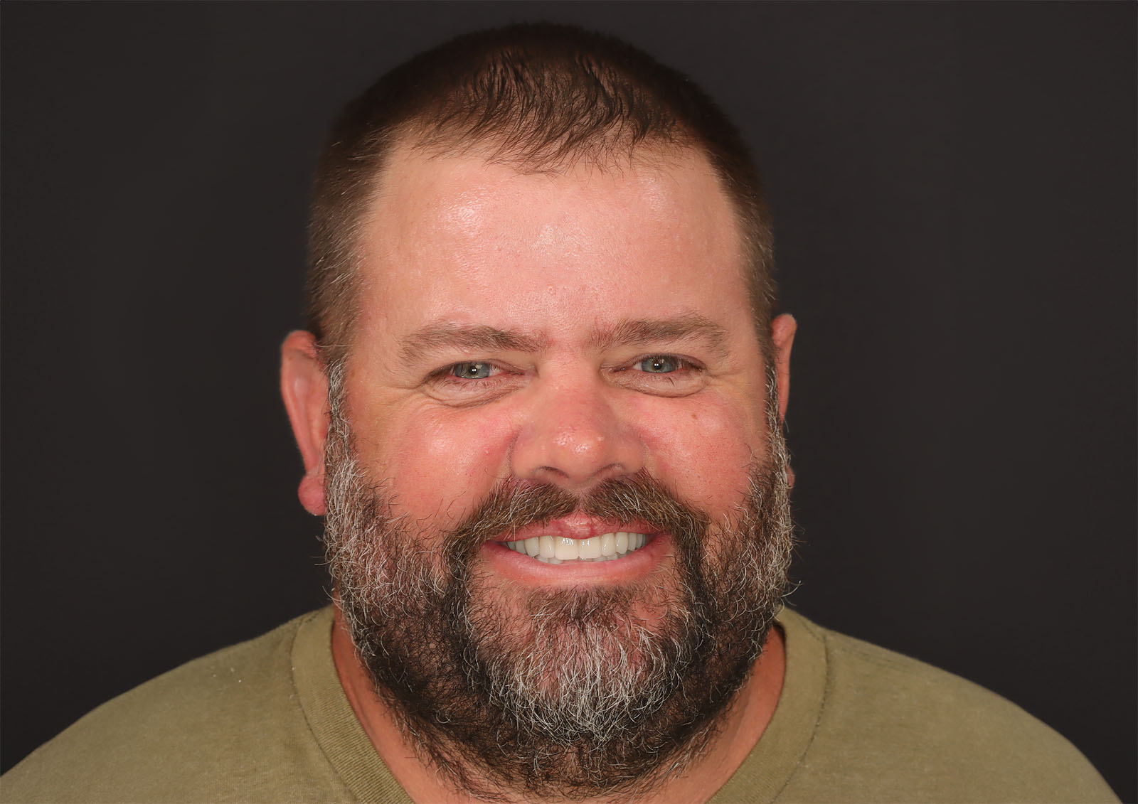 A bearded man with short dark hair wearing a green T-shirt smiles at the camera against a black background.