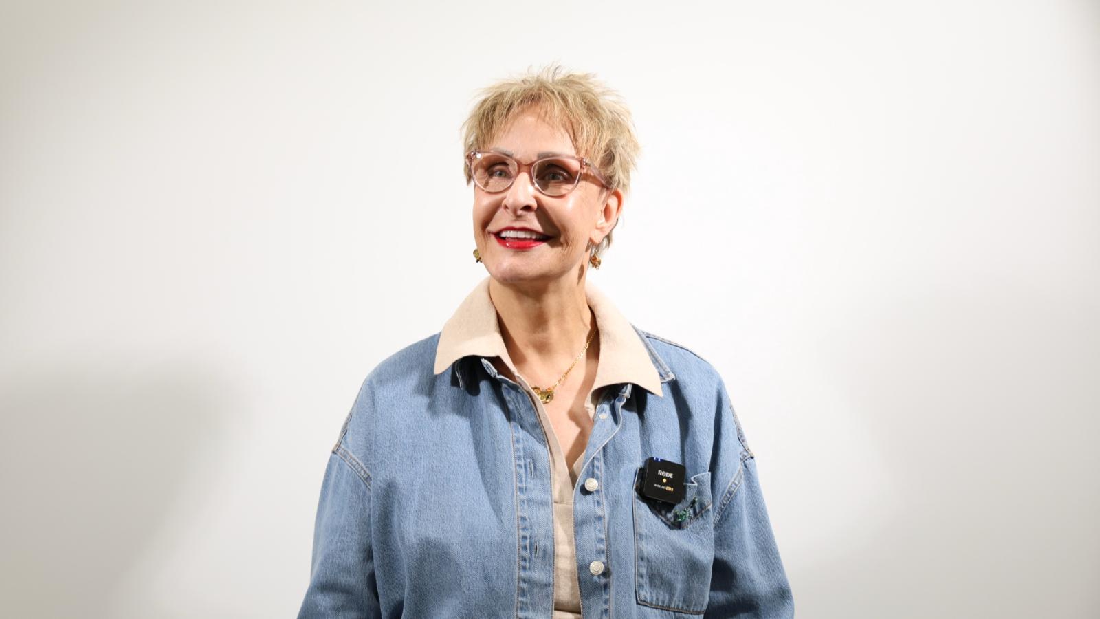 A woman with short blond hair and glasses wearing a denim jacket and beige shirt stands smiling against a plain white background.