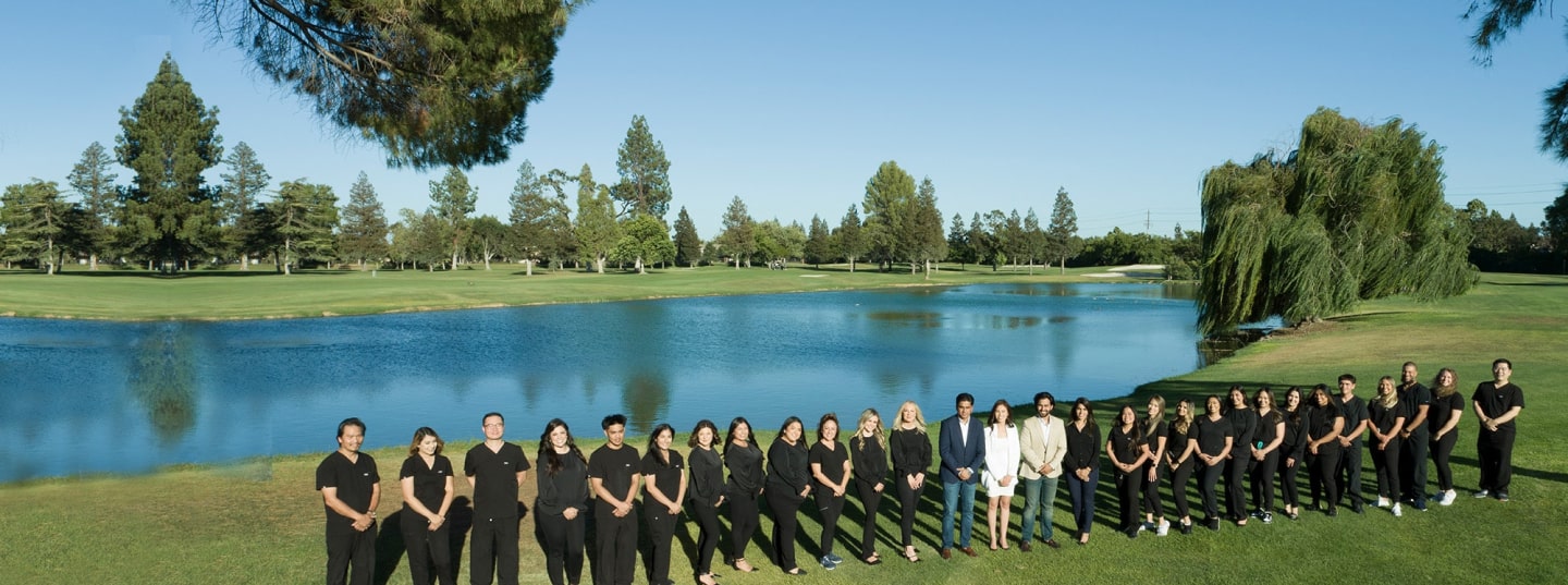 A group of people stands in a line on grassy ground near a lake, with trees and a clear sky in the background.